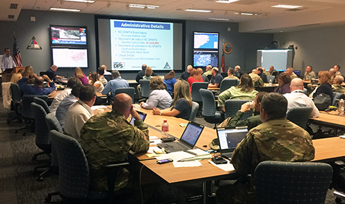 National Guard members and other disaster relief staff work together to coordinate emergency relief efforts in the aftermath of Hurricane Matthew. (Volpe photo) A large conference room is filled with National Guard members and other emergency response staff who are working on laptops while also viewing a presentation at the front of the room.