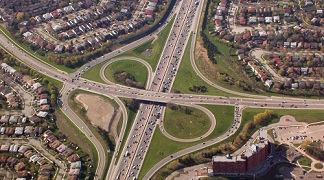 A twisted highway junction surrounded on the outskirts by housing complexes and a large building.