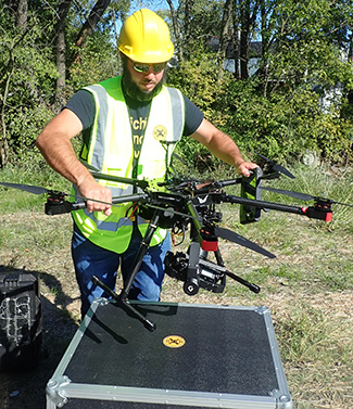 A man wearing a hard hat and safety vest holds a drone that's equipped with a camera.