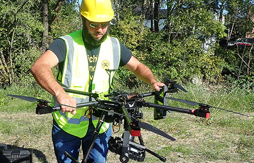 A man wearing a hard hat and safety vest holds a drone that's equipped with a camera.