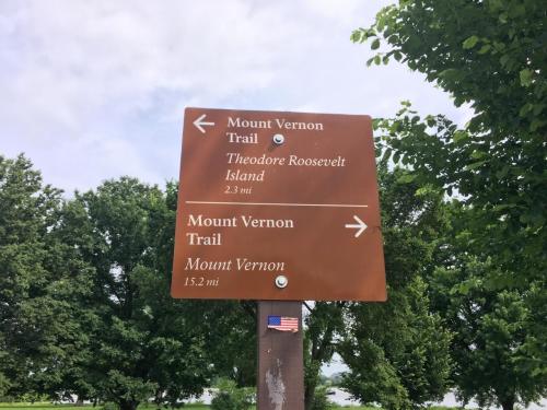 "Brown bicycle-pedestrian path sign that says 'Mount Vernon Trail, Theodore Roosevelt Island 2.3 miles' with an arrow pointing left. On the bottom of the sign is an arrow pointing write with the text 'Mount Vernon 15.2 miles."