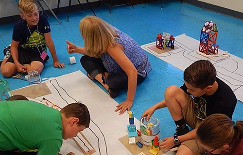 A teacher and several students sit on the floor testing small robotic vehicles with marker-drawn roads on large sheets of paper.