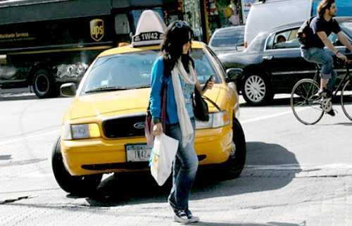 A woman crosses the street in front of an approaching taxi cab while looking down at her cell phone.