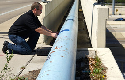 A man kneels down, testing a pipeline that runs along a bridge overpass.