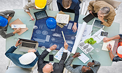 Image of construction workers with their hats on gathering around a plan, spread across a table
