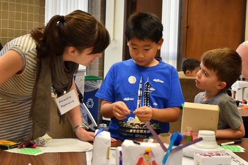 A Volpe transportation planner helps two small children assemble mock cities using cardboard blocks, markers, glue, stickers, and construction paper.