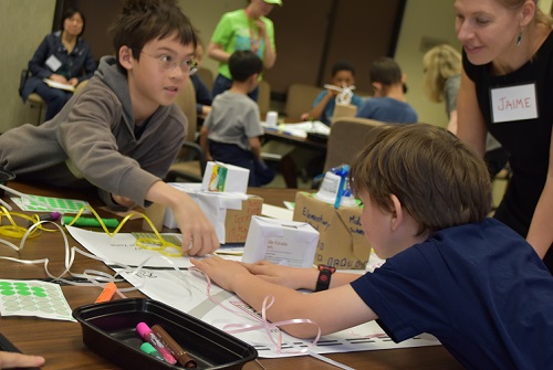 A Volpe transportation planner helps two small children assemble mock cities using cardboard blocks, markers, glue, stickers, ribbon, and construction paper.