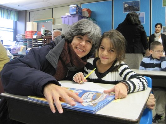A woman and a student smile together from the student's school desk during a reading session.