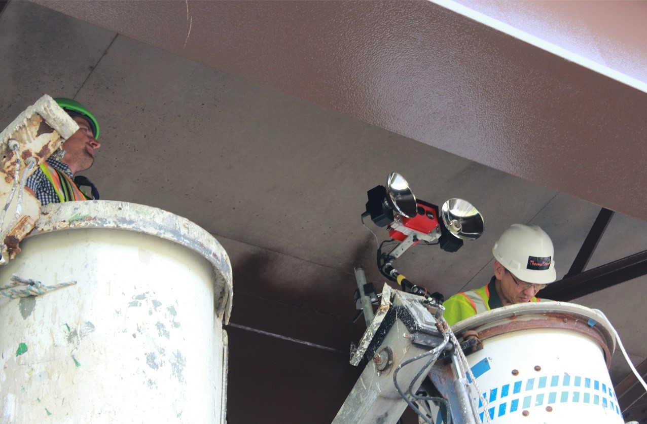 Two people in hard hats in truck buckets measuring paint on a steel bridge