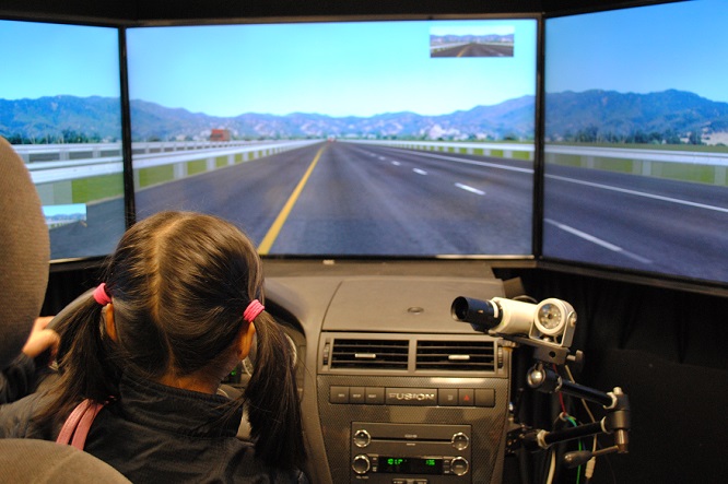 A girl sits in the seat of Volpe's car simulator with her hands on the wheel, navigating through a driving scenario.