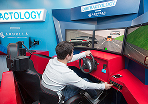 A man sit in the seat of a driving simulator with a sign above him that reads "distractology." The simulator is Arbella branded.