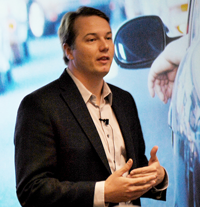 Chris Urmson speaks at Volpe in front of a screen that shows a car in traffic.