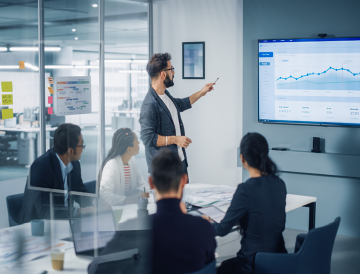 A man standing in front of a boardroom presenting slides on a television