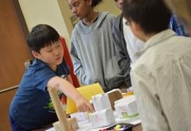 A child points out various elements of the city he created using paper boxes and markers.