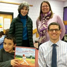 An elementary school student holds up a book as he sits beside his Volpe Lunch Buddy and two school staff members.