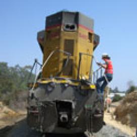An engineer inspects a train that was damaged in a crash. Photo of a stationary train with a railroad employee climbing down the train's side-deck.